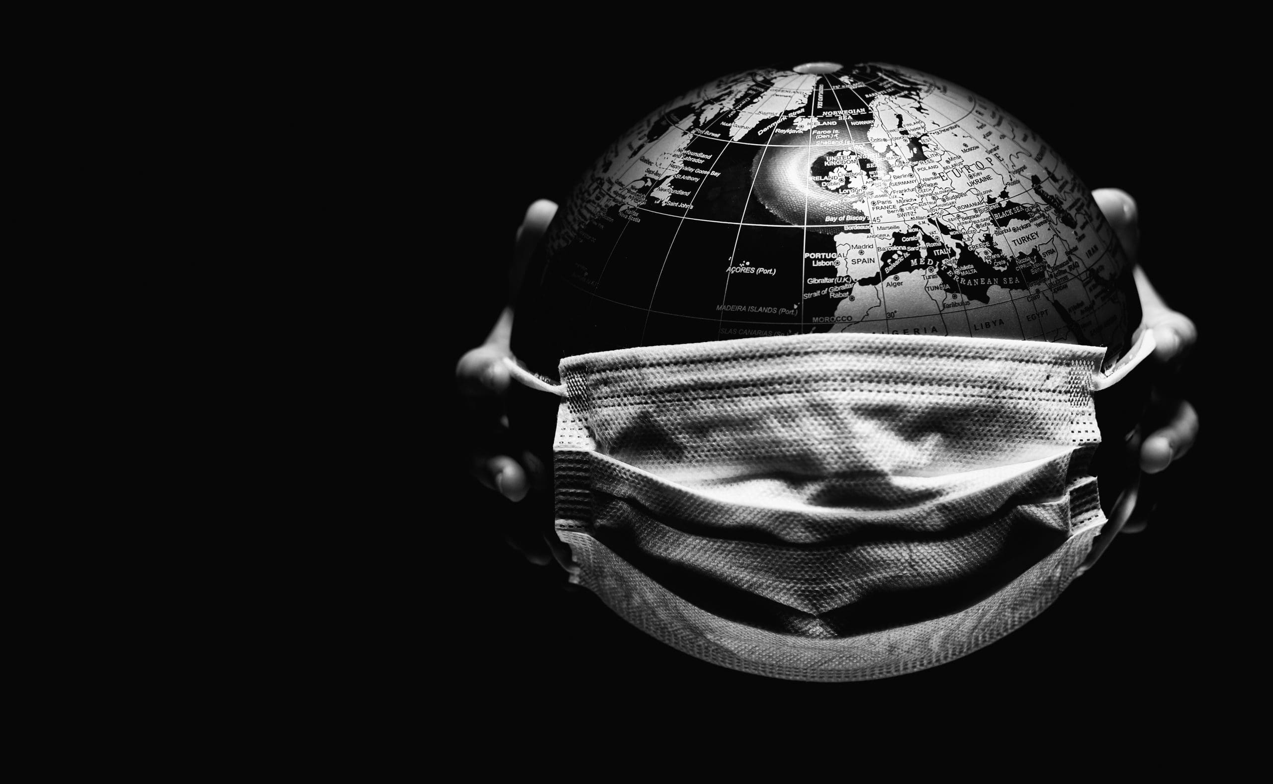 Hands of little child holding globe sphere, planet map covered with medical protective mask isolated on black background. Concept of COVID-19 pandemic infection