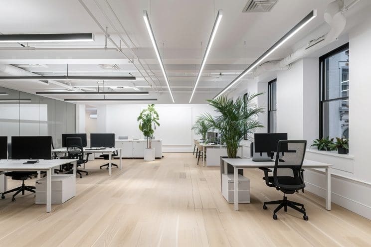 A white office with modern desks and chairs, with LED lights on the ceiling illuminating an empty wall for text or artwork. The spacious and clean space