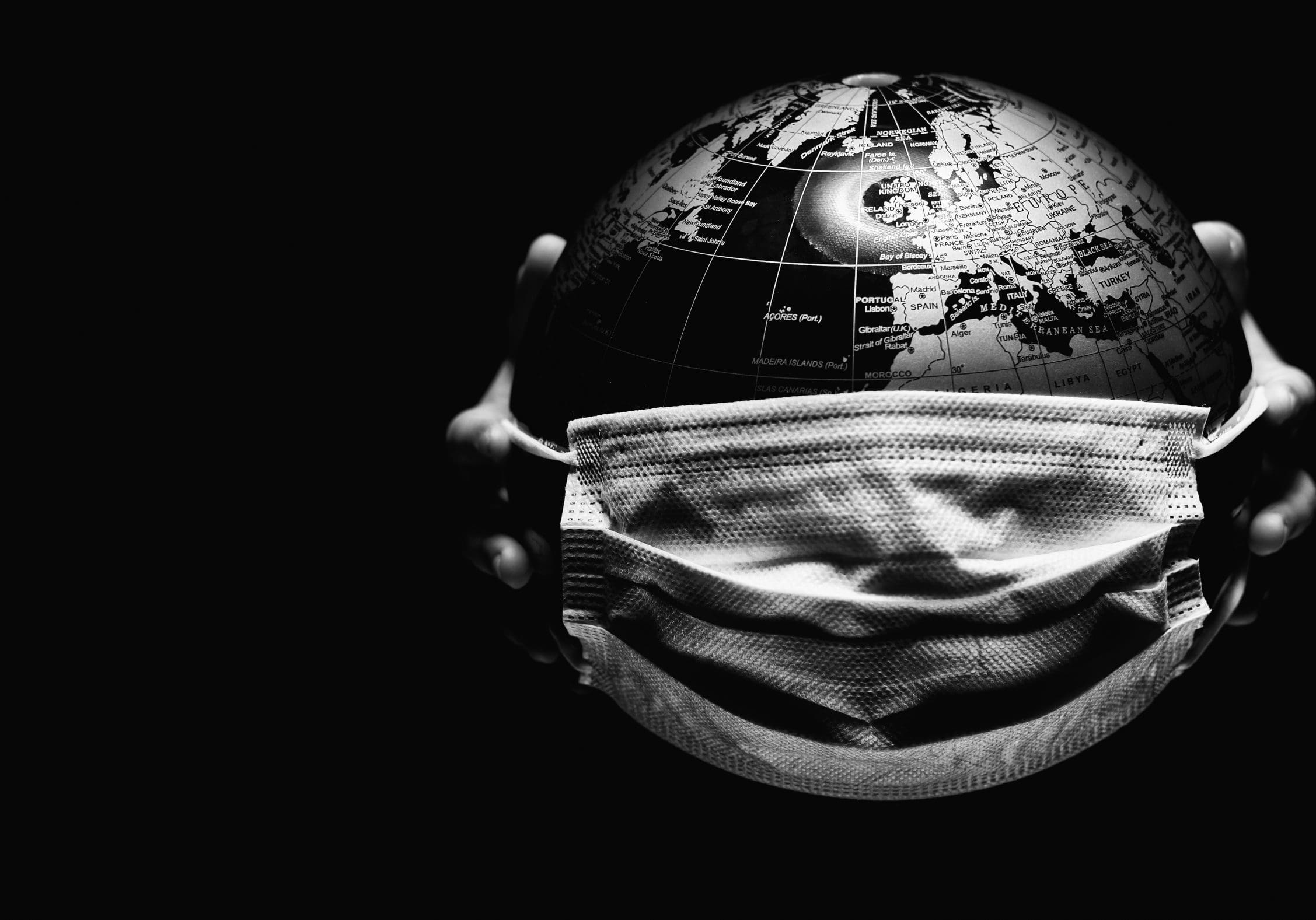 Hands of little child holding globe sphere, planet map covered with medical protective mask isolated on black background. Concept of COVID-19 pandemic infection